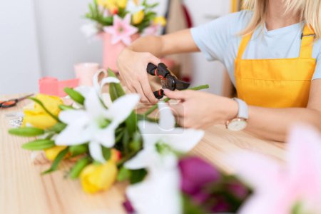Téléchargez les photos : Young blonde woman florist cutting bouquet of flowers at flower shop - en image libre de droit