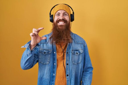 Photo for Caucasian man with long beard listening to music using headphones smiling and confident gesturing with hand doing small size sign with fingers looking and the camera. measure concept. - Royalty Free Image