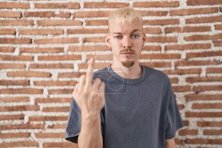 Foto de Young caucasian man standing over bricks wall showing middle finger, impolite and rude fuck off expression - Imagen libre de derechos