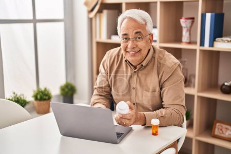 Téléchargez les photos : Senior man using laptop holding pills at home - en image libre de droit