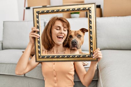 Photo for Young caucasian woman holding empty frame sitting on floor with dog at home - Royalty Free Image