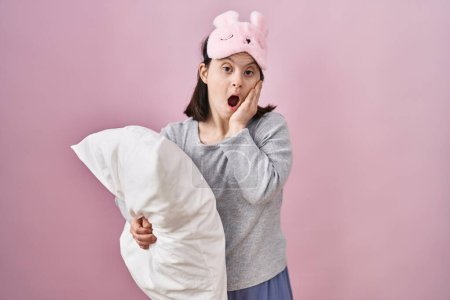 Téléchargez les photos : Woman with down syndrome wearing sleeping mask hugging pillow afraid and shocked, surprise and amazed expression with hands on face - en image libre de droit