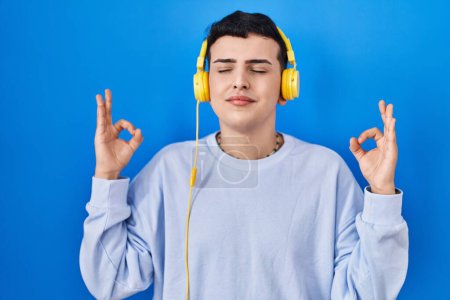 Téléchargez les photos : Personne non binaire écoutant de la musique à l'aide d'écouteurs détendus et souriants avec les yeux fermés faisant geste de méditation avec les doigts. concept de yoga. - en image libre de droit