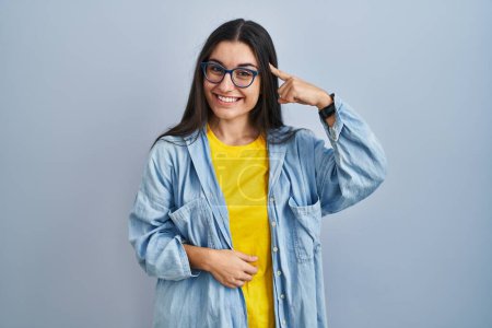 Foto de Young hispanic woman standing over blue background smiling pointing to head with one finger, great idea or thought, good memory - Imagen libre de derechos