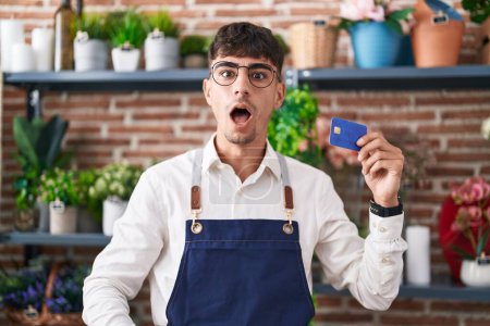 Téléchargez les photos : Jeune homme hispanique travaillant au magasin de fleuriste tenant la carte de crédit effrayé et étonné avec la bouche ouverte pour la surprise, visage d'incrédulité - en image libre de droit