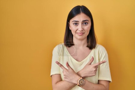 Téléchargez les photos : Hispanic girl wearing casual t shirt over yellow background pointing to both sides with fingers, different direction disagree - en image libre de droit