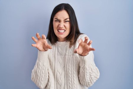 Téléchargez les photos : Jeune femme brune debout sur fond bleu souriant drôle faisant griffe geste comme chat, expression agressive et sexy - en image libre de droit