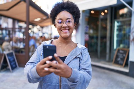 Foto de Mujer afroamericana sonriendo confiada usando teléfono inteligente en la terraza de la cafetería - Imagen libre de derechos