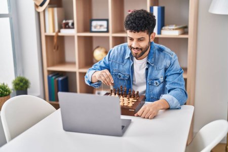 Téléchargez les photos : Jeune homme arabe ayant jeu d'échecs en ligne assis sur la table à la maison - en image libre de droit
