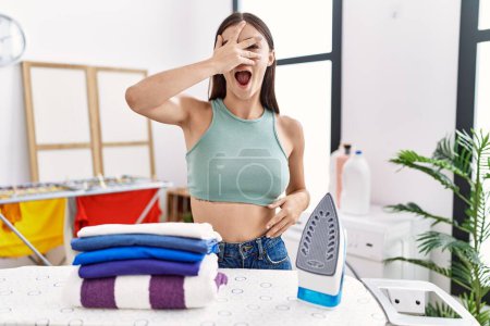 Photo for Young hispanic woman ironing clothes at laundry room peeking in shock covering face and eyes with hand, looking through fingers with embarrassed expression. - Royalty Free Image