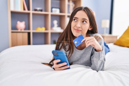 Foto de Joven mujer hispana hermosa usando teléfono inteligente y tarjeta de crédito tumbado en la cama en el dormitorio - Imagen libre de derechos
