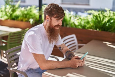 Foto de Joven pelirrojo usando teléfono inteligente sentado en la mesa en la terraza de la cafetería - Imagen libre de derechos