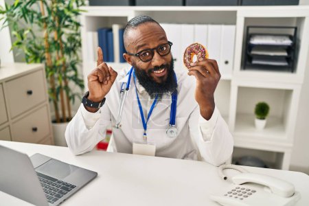 Foto de African american man working at dietitian clinic holding doughnut surprised with an idea or question pointing finger with happy face, number one - Imagen libre de derechos