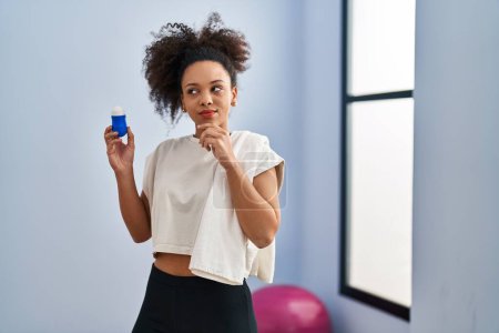 Foto de Young african american woman wearing sportswear and towel holding deodorant serious face thinking about question with hand on chin, thoughtful about confusing idea - Imagen libre de derechos