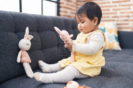 Foto de Adorable bebé hispano sosteniendo juguete sentado en el sofá en casa - Imagen libre de derechos