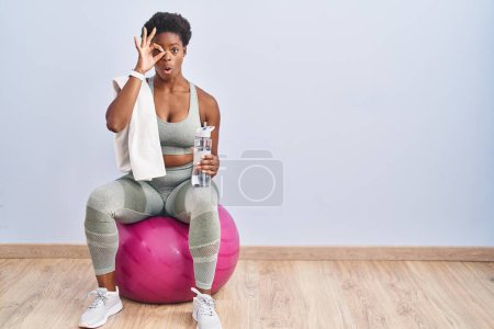 Foto de Mujer afroamericana usando ropa deportiva sentada en la pelota de pilates haciendo un gesto bien sorprendido con la cara sorprendida, los ojos mirando a través de los dedos. expresión incrédula. - Imagen libre de derechos