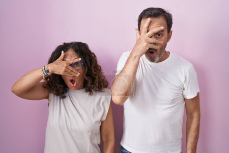 Photo for Middle age hispanic couple together over pink background peeking in shock covering face and eyes with hand, looking through fingers with embarrassed expression. - Royalty Free Image