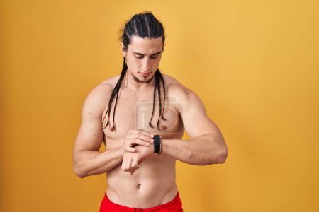 Photo for Hispanic man with long hair standing shirtless over yellow background checking the time on wrist watch, relaxed and confident - Royalty Free Image