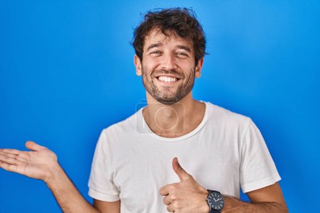 Foto de Hispanic young man standing over blue background showing palm hand and doing ok gesture with thumbs up, smiling happy and cheerful - Imagen libre de derechos