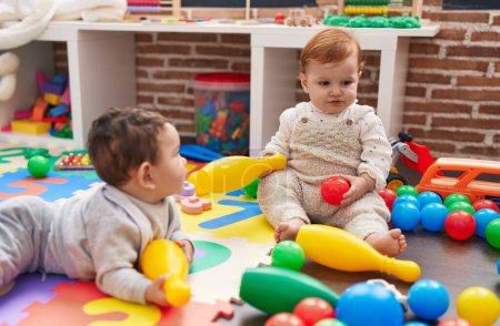 Foto de Dos adorables bebés jugando con pelotas y bolos sentados en el suelo en el jardín de infantes - Imagen libre de derechos