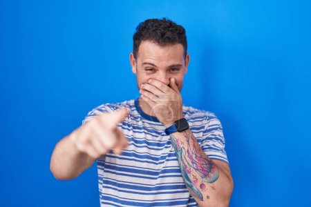 Photo for Young hispanic man standing over blue background laughing at you, pointing finger to the camera with hand over mouth, shame expression - Royalty Free Image