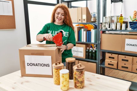Téléchargez les photos : Jeune rousse femme portant l'uniforme volontaire emballage boîte de dons au centre de charité - en image libre de droit