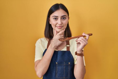 Photo for Hispanic girl eating healthy  wooden spoon looking confident at the camera smiling with crossed arms and hand raised on chin. thinking positive. - Royalty Free Image