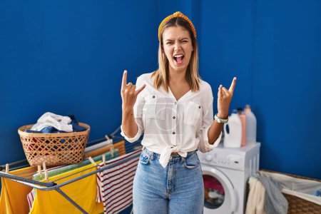 Téléchargez les photos : Young blonde woman at laundry room shouting with crazy expression doing rock symbol with hands up. music star. heavy music concept. - en image libre de droit