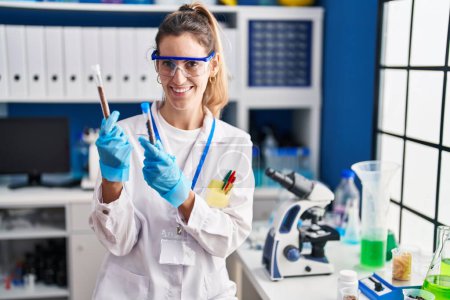 Foto de Mujer joven científica sonriendo confiada sosteniendo tubos de ensayo en el laboratorio - Imagen libre de derechos