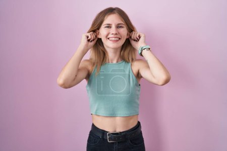 Photo for Blonde caucasian woman standing over pink background smiling pulling ears with fingers, funny gesture. audition problem - Royalty Free Image