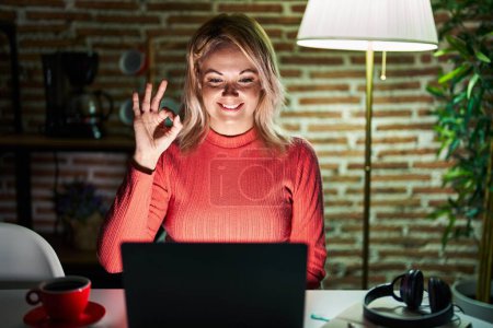 Photo for Blonde woman using laptop at night at home smiling positive doing ok sign with hand and fingers. successful expression. - Royalty Free Image
