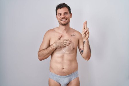 Téléchargez les photos : Young hispanic man standing shirtless wearing underware smiling swearing with hand on chest and fingers up, making a loyalty promise oath - en image libre de droit