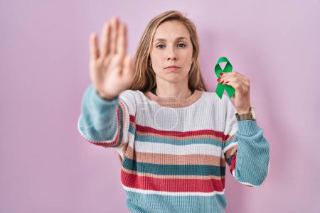 Photo for Young blonde woman holding support green ribbon with open hand doing stop sign with serious and confident expression, defense gesture - Royalty Free Image