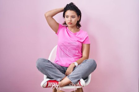 Foto de Hispanic young woman sitting on chair over pink background confuse and wondering about question. uncertain with doubt, thinking with hand on head. pensive concept. - Imagen libre de derechos