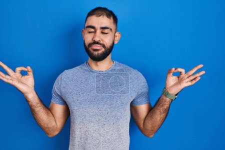 Téléchargez les photos : Homme du Moyen-Orient avec la barbe debout sur fond bleu détendu et souriant avec les yeux fermés faisant geste de méditation avec les doigts. concept de yoga. - en image libre de droit
