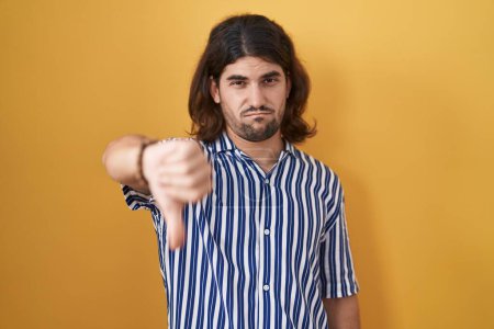 Foto de Hispanic man with long hair standing over yellow background looking unhappy and angry showing rejection and negative with thumbs down gesture. bad expression. - Imagen libre de derechos