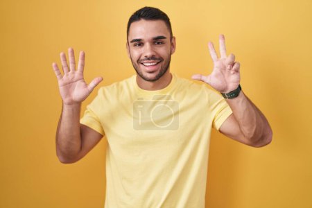 Photo for Young hispanic man standing over yellow background showing and pointing up with fingers number eight while smiling confident and happy. - Royalty Free Image