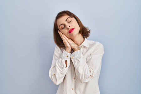 Foto de Young beautiful woman standing casual over blue background sleeping tired dreaming and posing with hands together while smiling with closed eyes. - Imagen libre de derechos