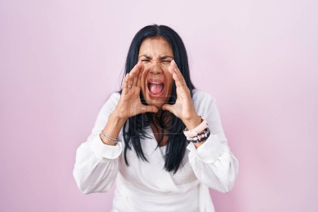 Photo for Mature hispanic woman standing over pink background shouting angry out loud with hands over mouth - Royalty Free Image