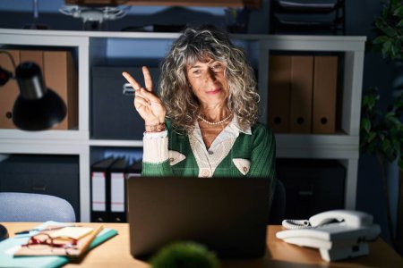 Photo for Middle age woman working at night using computer laptop smiling with happy face winking at the camera doing victory sign with fingers. number two. - Royalty Free Image
