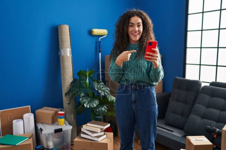Téléchargez les photos : Young hispanic woman at new home using smartphone smiling happy pointing with hand and finger - en image libre de droit