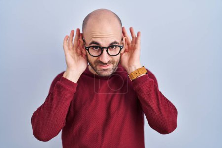 Foto de Young bald man with beard standing over white background wearing glasses trying to hear both hands on ear gesture, curious for gossip. hearing problem, deaf - Imagen libre de derechos