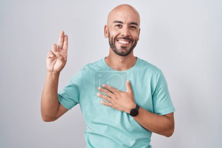 Foto de Hombre calvo de mediana edad de pie sobre fondo blanco sonriendo jurando con la mano en el pecho y los dedos hacia arriba, haciendo una promesa de lealtad juramento - Imagen libre de derechos