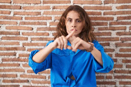 Foto de Beautiful brunette woman standing over bricks wall rejection expression crossing fingers doing negative sign - Imagen libre de derechos