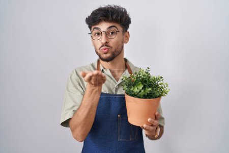Foto de Arab man with beard holding green plant pot looking at the camera blowing a kiss with hand on air being lovely and sexy. love expression. - Imagen libre de derechos