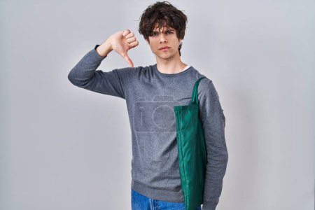 Foto de Young man wearing reusable bag with angry face, negative sign showing dislike with thumbs down, rejection concept - Imagen libre de derechos