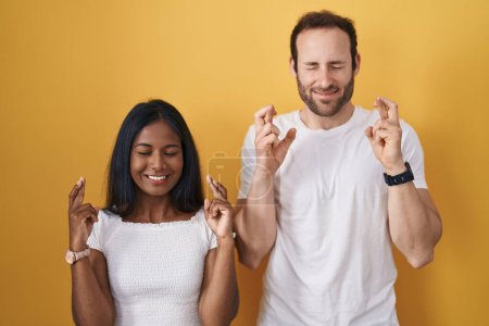 Téléchargez les photos : Interracial couple standing over yellow background gesturing finger crossed smiling with hope and eyes closed. luck and superstitious concept. - en image libre de droit