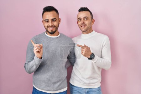 Téléchargez les photos : Couple homosexuel debout sur fond rose avec un grand sourire sur le visage, pointant avec le doigt de la main sur le côté en regardant la caméra. - en image libre de droit
