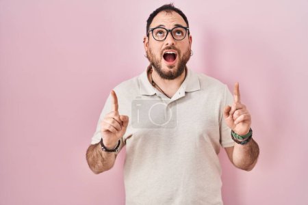 Téléchargez les photos : Plus size hispanic man with beard standing over pink background amazed and surprised looking up and pointing with fingers and raised arms. - en image libre de droit