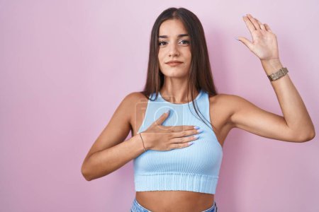 Photo for Young brunette woman standing over pink background swearing with hand on chest and open palm, making a loyalty promise oath - Royalty Free Image